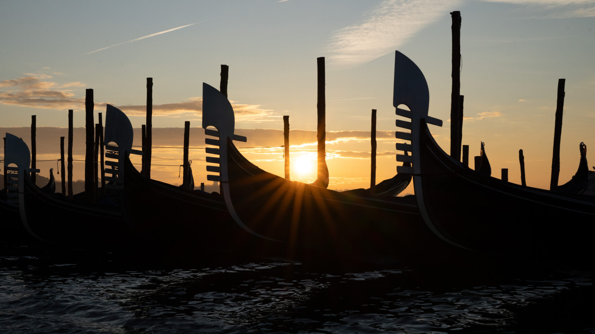 - The Travel Captains Silhouetted Venetian gondolas docked along a canal at sunset with tall mooring poles against an orange sky