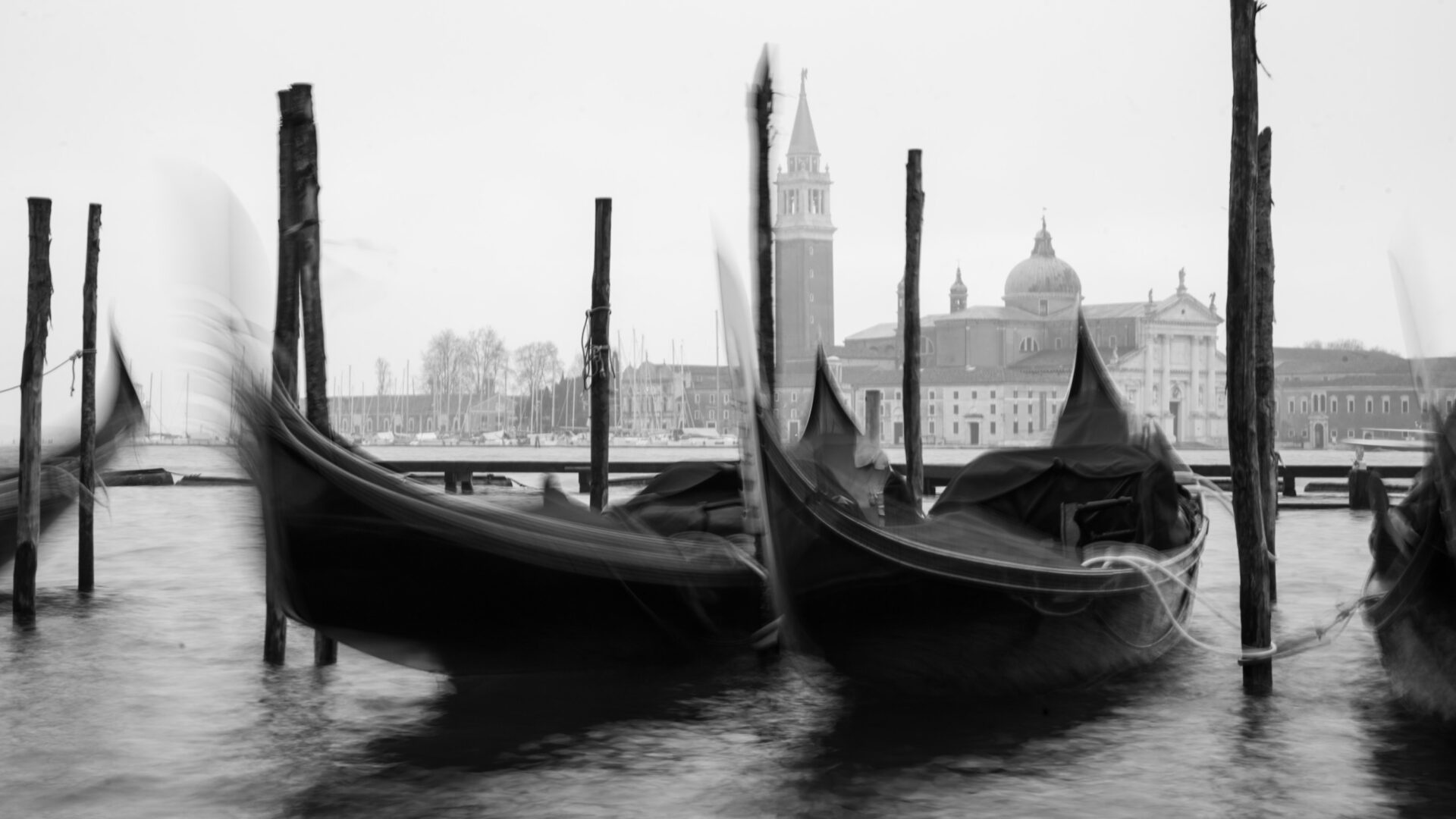 - The Travel Captains Gondolas tied to wooden posts on a Venetian canal with historic buildings in the hazy background.