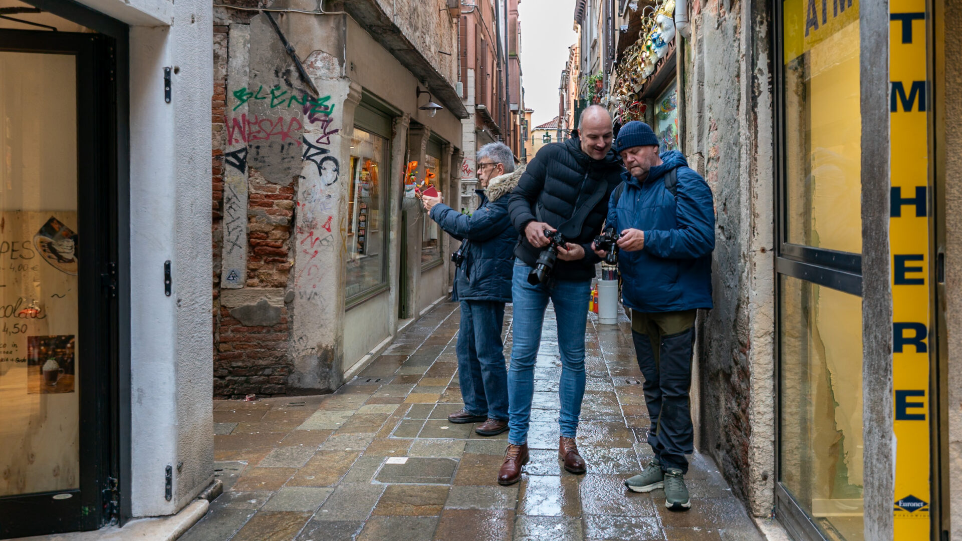 - The Travel Captains Three men with cameras stand in a narrow, graffiti-covered alley, examining their gear on a wet, tiled pavement, with shopfronts on both sides.